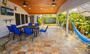 Eine überdachte Terrasse mit Esstisch und Stühlen, einem an der Wand befestigten Fernseher und einer Hängematte.