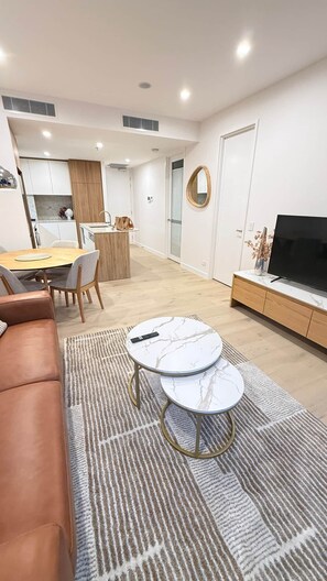 Ein modernes Wohnzimmer mit einem Ledersofa, einem Fernseher auf einer Marmorkonsole und einem Essbereich mit Holztisch und Stühlen.