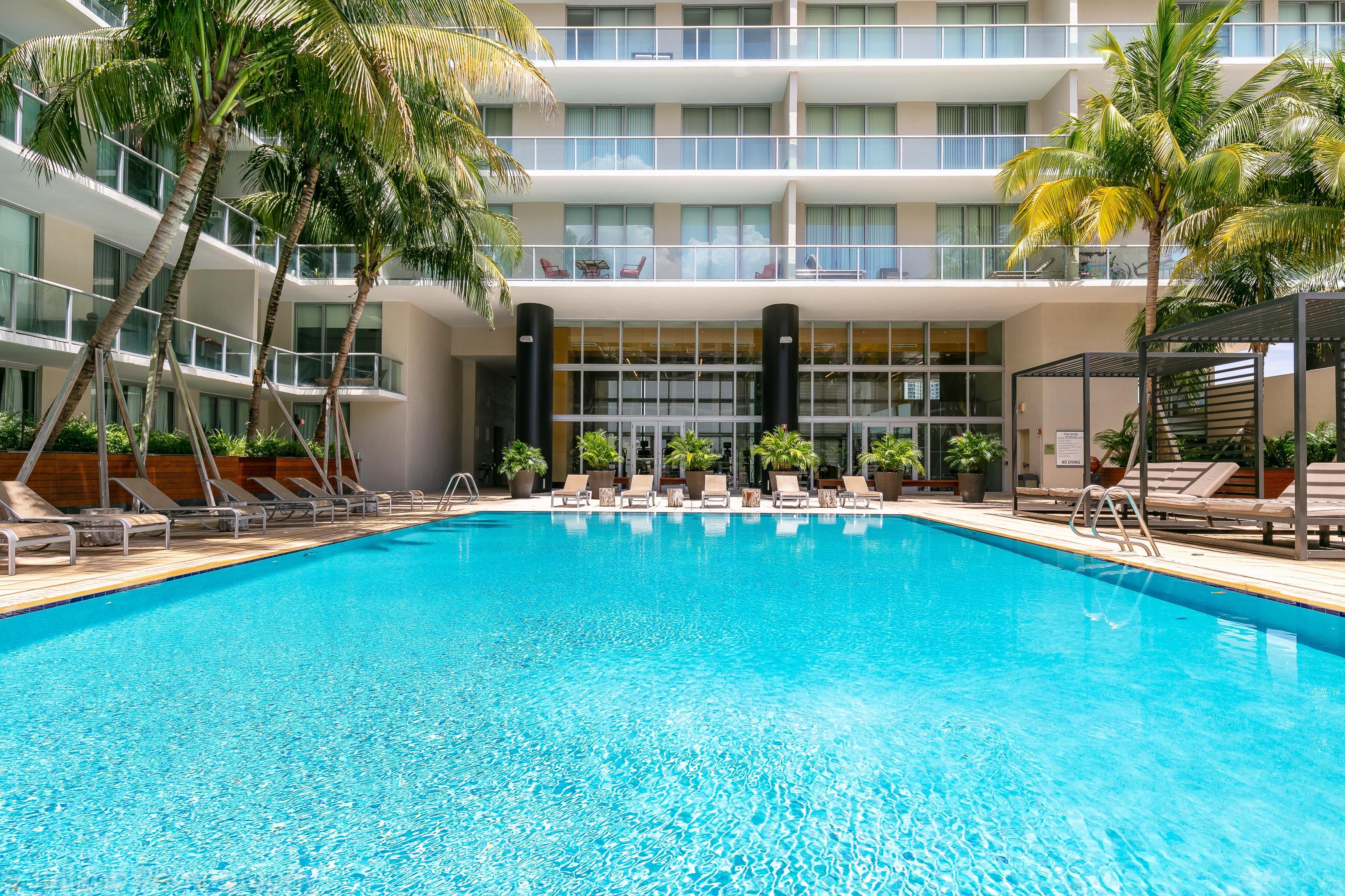 A swimming pool with lounge chairs and palm trees, surrounded by a multi-story building with balconies.