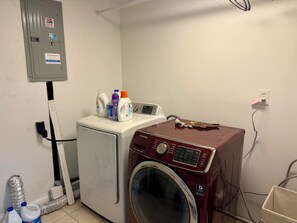 Uma lavanderia com máquina de lavar e secadora, medidor de consumo e produtos de limpeza em cima da secadora.