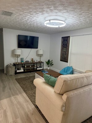 A living room with a beige sofa, a coffee table, a TV mounted on the wall, a lamp, and a decorative wall piece. - 3 BR waterfront house 25 minutes from TAMPA!! Feat. Pool Kayaks, FIshing & Dock! (Ruskin)