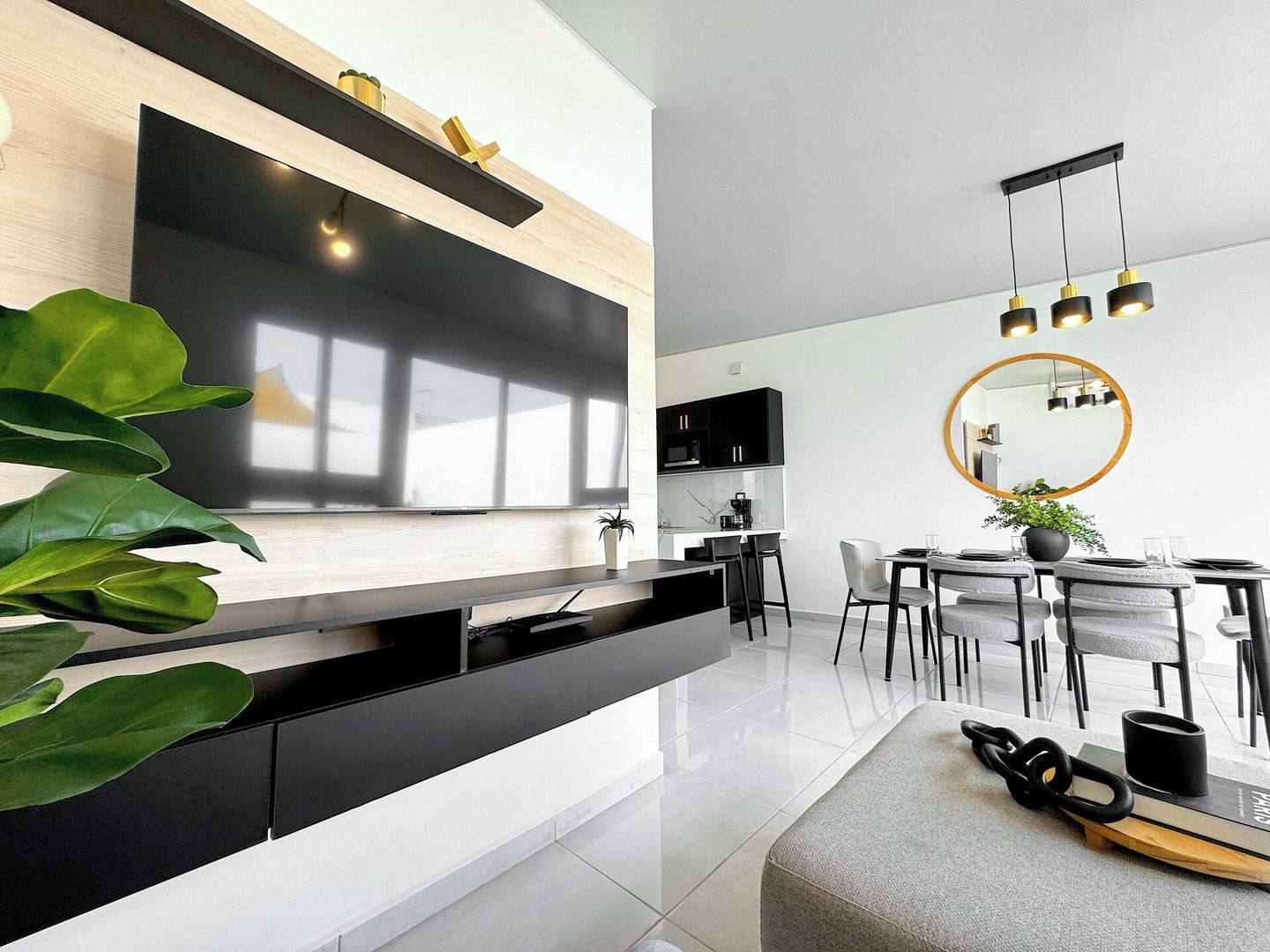 A modern kitchen with a large flat-screen TV, black cabinetry, and a dining area with a round mirror and pendant lights.