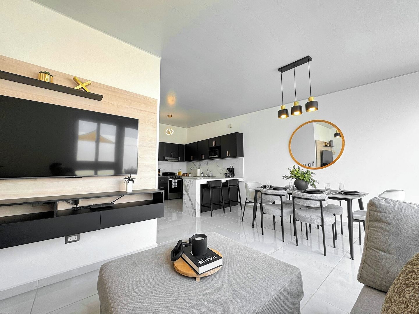 A modern living room with a flat-screen TV, a dining area with a round table and chairs, and a kitchen with dark cabinets and a marble countertop.