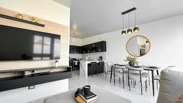 A modern living room with a flat-screen TV, a dining area with a round table and chairs, and a kitchen with dark cabinets and a marble countertop.