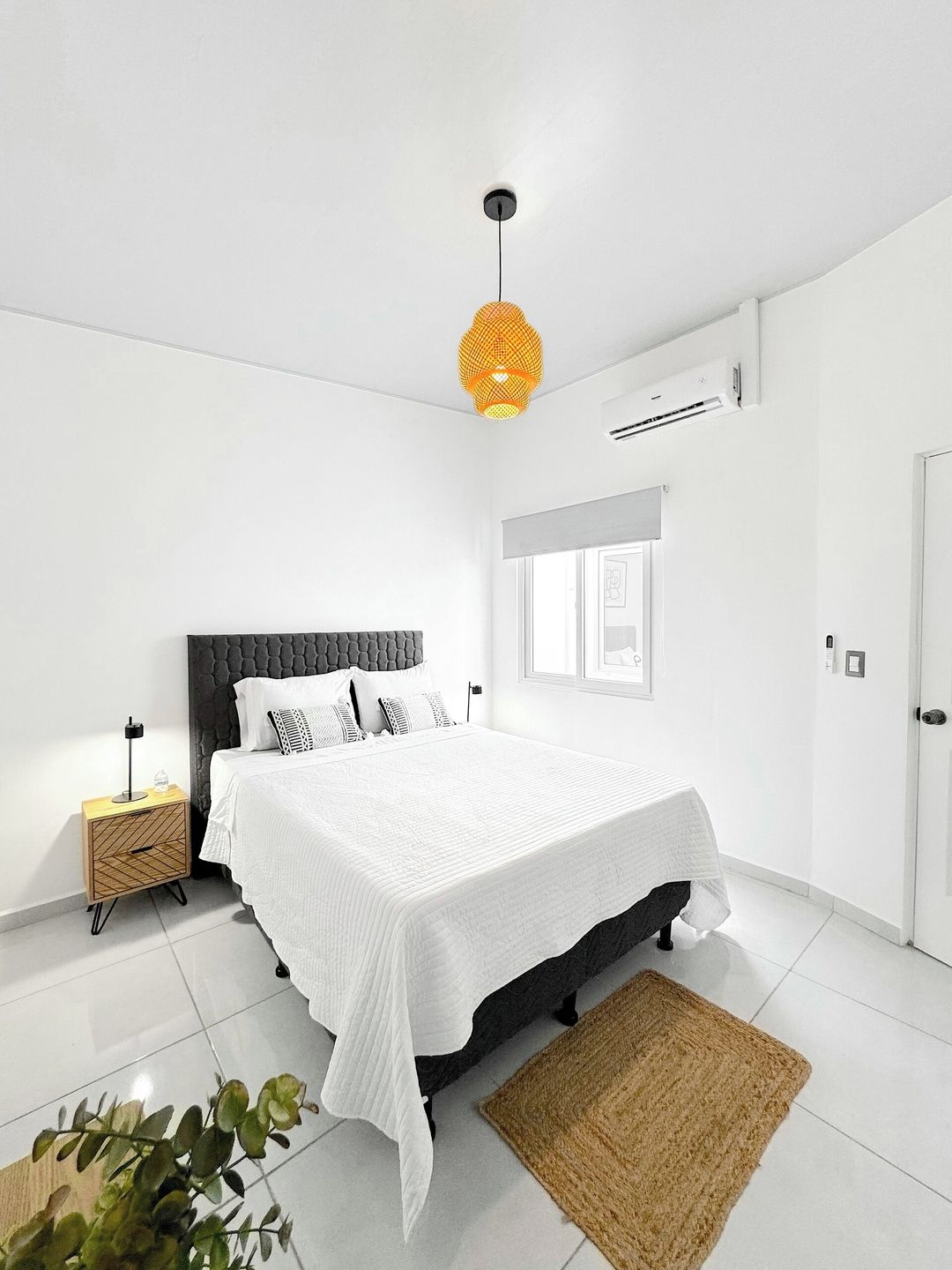 A modern bedroom with a large bed, a bedside table, an air conditioning unit, and a hanging light fixture.