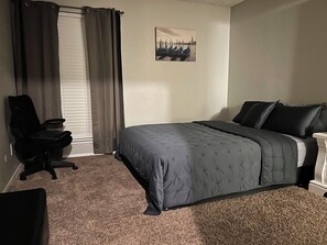 Una camera da letto con un letto, una sedia, una finestra con tende e un quadro appeso al muro.