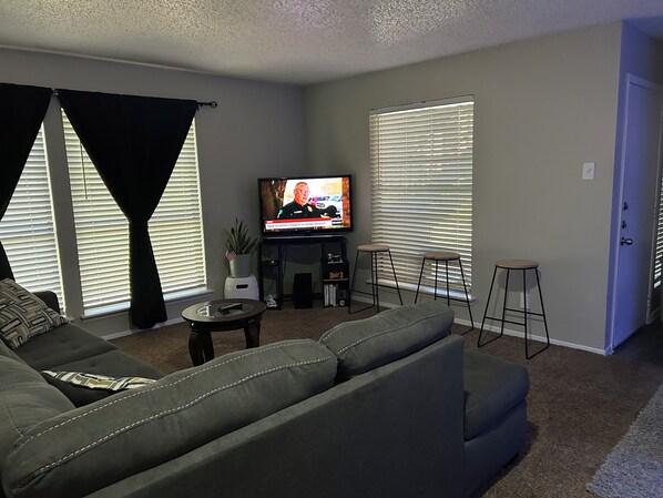 A living room with a large flat-screen TV, a sofa, a coffee table, and two bar stools.