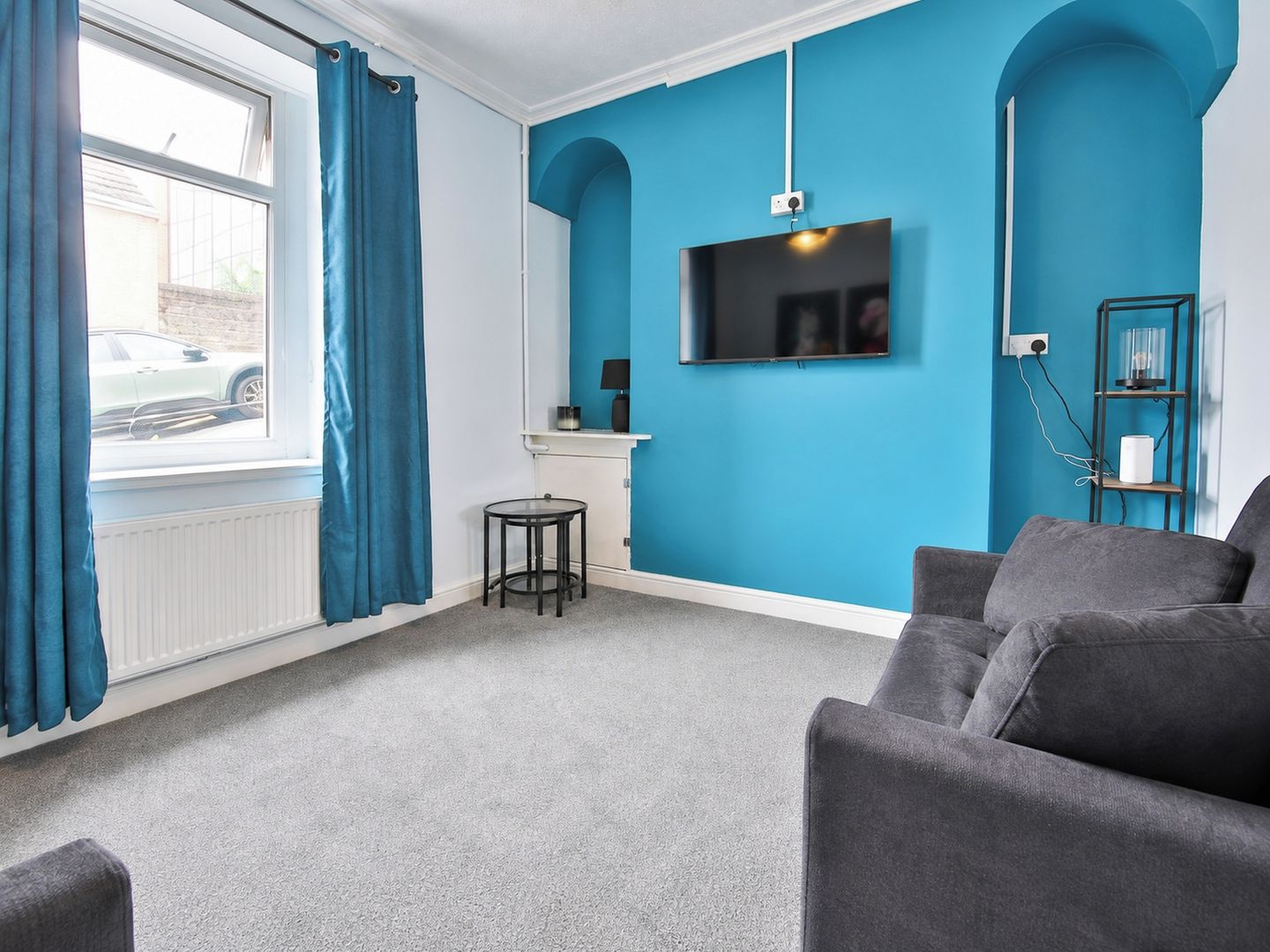 A room with a grey sofa, a small round table, a TV mounted on a blue wall, and a window with curtains.
