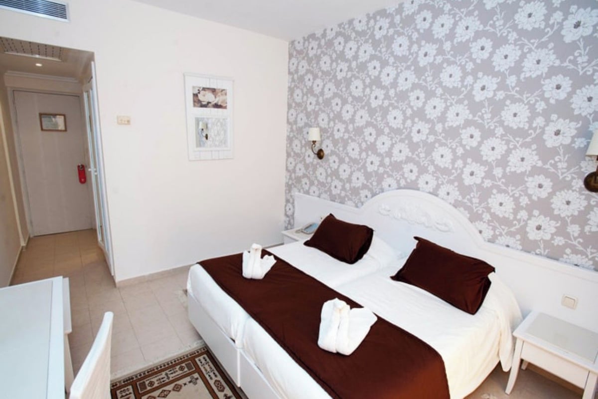 A double bed with a brown bedspread and two pillows, a white nightstand, a wall with a floral pattern, and a door with a red handle.