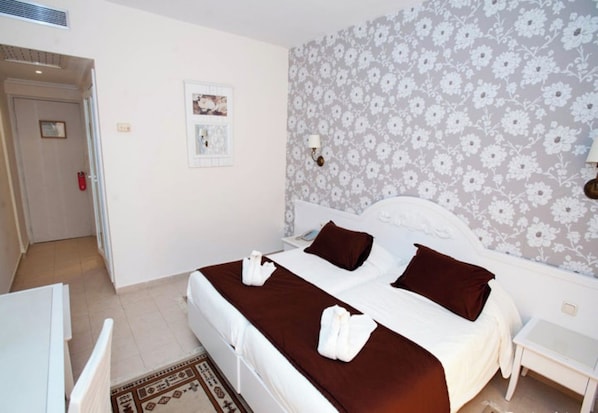 A double bed with a brown bedspread and two pillows, a white nightstand, a wall with a floral pattern, and a door with a red handle.