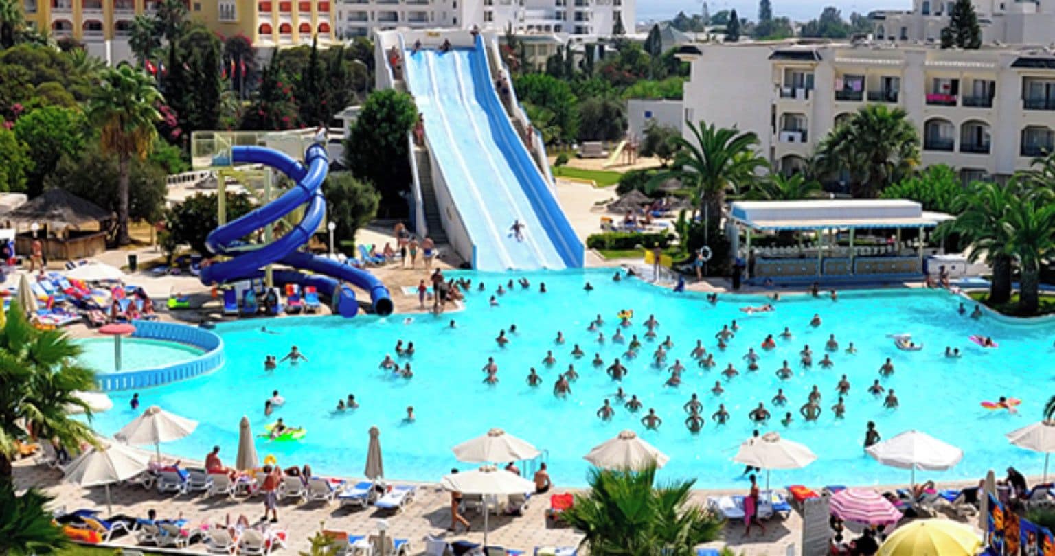 A large swimming pool with multiple slides and a busier area with umbrellas and lounge chairs.
