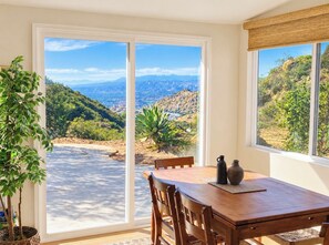 Un comedor con una mesa y sillas, vista a un paisaje montañoso a través de grandes puertas de vidrio y una planta en maceta a la izquierda.