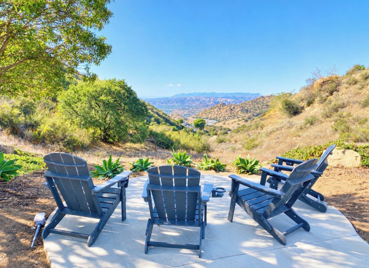 An outdoor seating area with Adirondack chairs and a table, surrounded by greenery and a view of a valley.