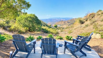 An outdoor seating area with Adirondack chairs and a table, surrounded by greenery and a view of a valley.