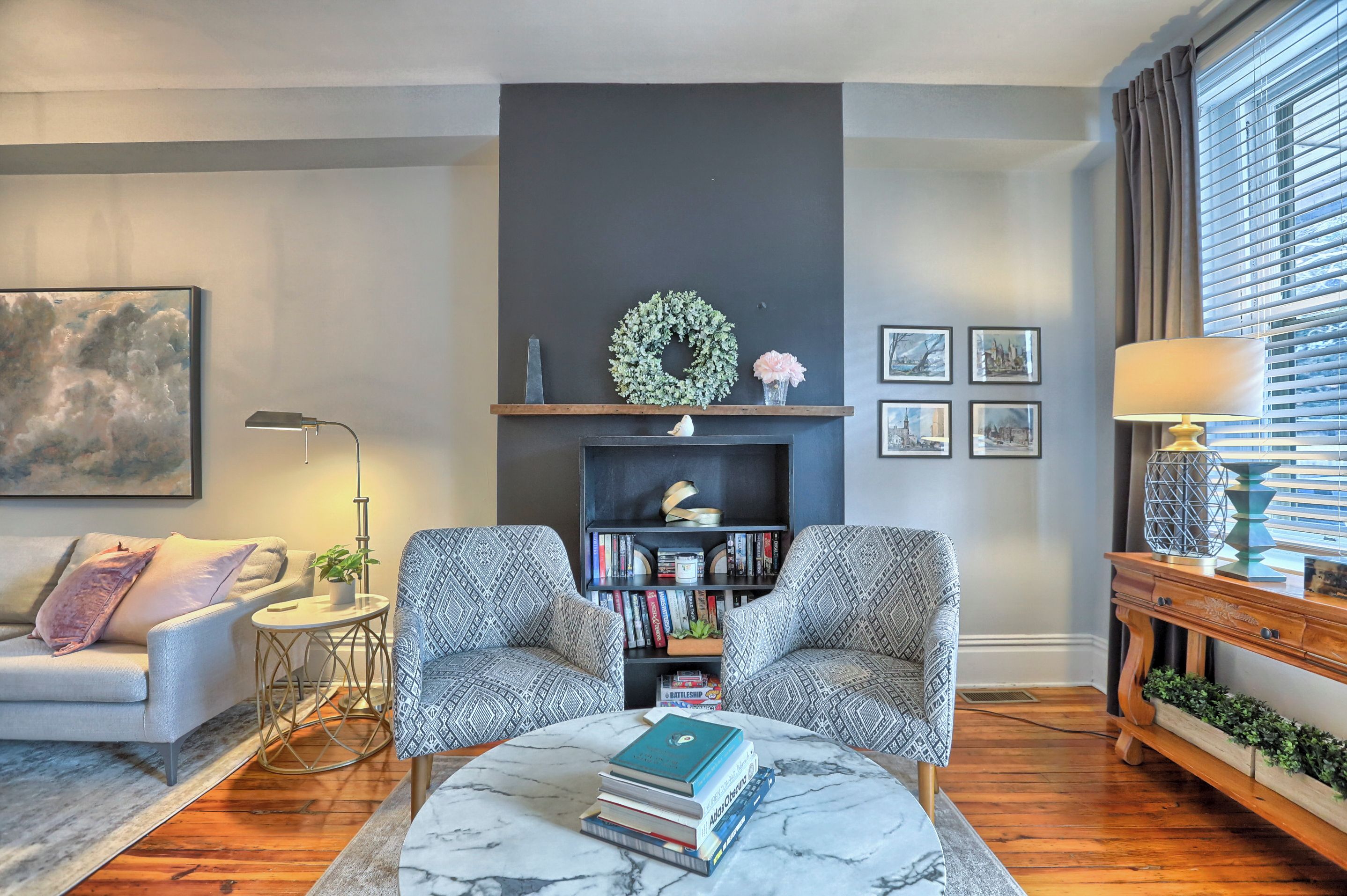 A modern living room with a sofa, two armchairs, a coffee table with books, a fireplace with a mantel, and a window with blinds.