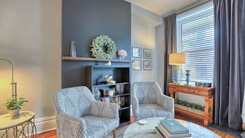 Ein Wohnzimmer mit zwei gemusterten Sesseln, einem Bücherregal, einem Couchtisch mit Büchern, einem Kamin und einem Fenster mit Vorhängen.