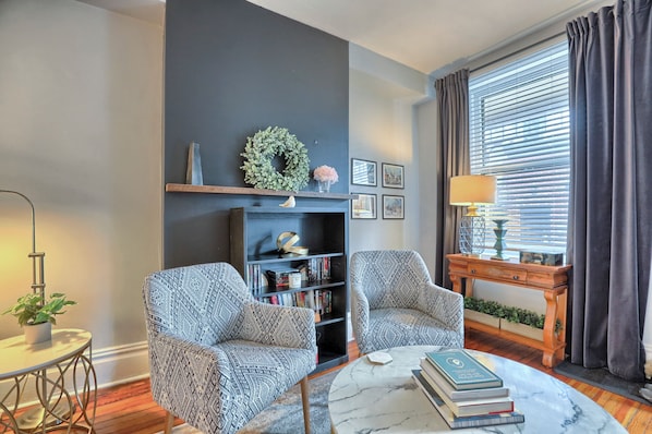 A living room with two patterned armchairs, a bookshelf, a coffee table with books, a fireplace, and a window with curtains.