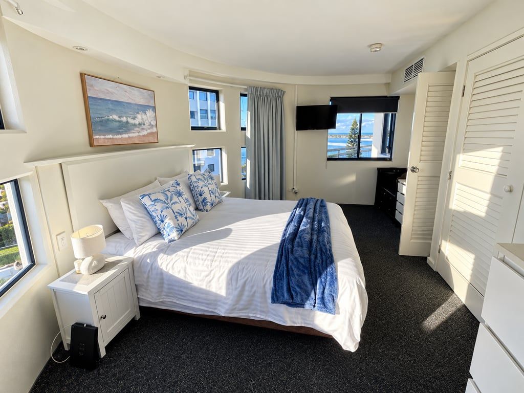 A hotel room with a bed, a bedside table with a lamp, a TV mounted on the wall, and a window with a view.