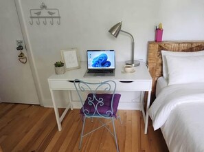 Ein weißer Schreibtisch mit einem Laptop, ein Metallstuhl, ein Nachttisch mit einer Lampe und ein Bett mit weißer Bettwäsche.