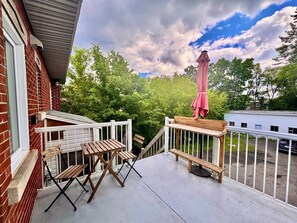 Ein Balkon mit einem Holztisch und Stühlen, einem rosa Sonnenschirm und Blick auf Bäume sowie ein Gebäude im Hintergrund.