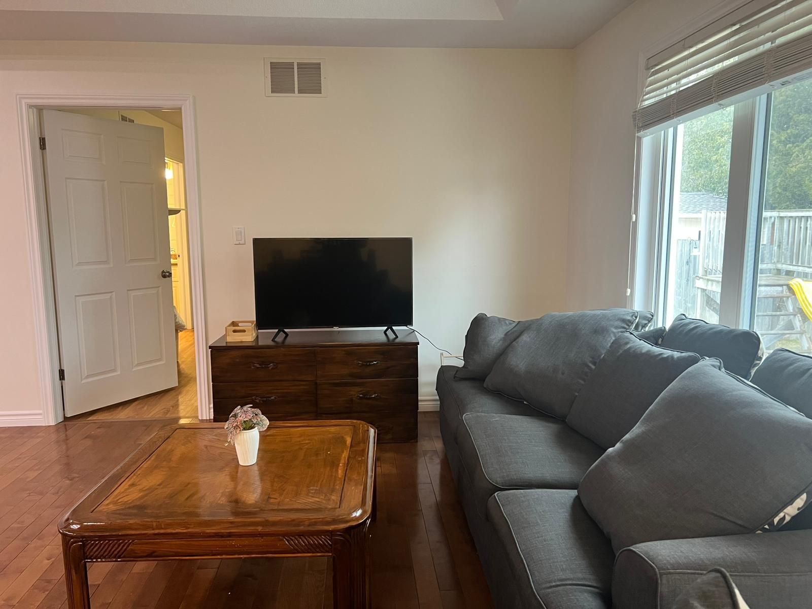 Ein Wohnzimmer mit Flachbildfernseher auf einer Holzkommode, einem grauen Sofa und einem Holztisch mit einer Vase.