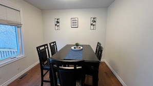 Een eetkamer met een rechthoekige tafel, zes stoelen, een plafondlamp en drie ingelijste foto's aan de muur.