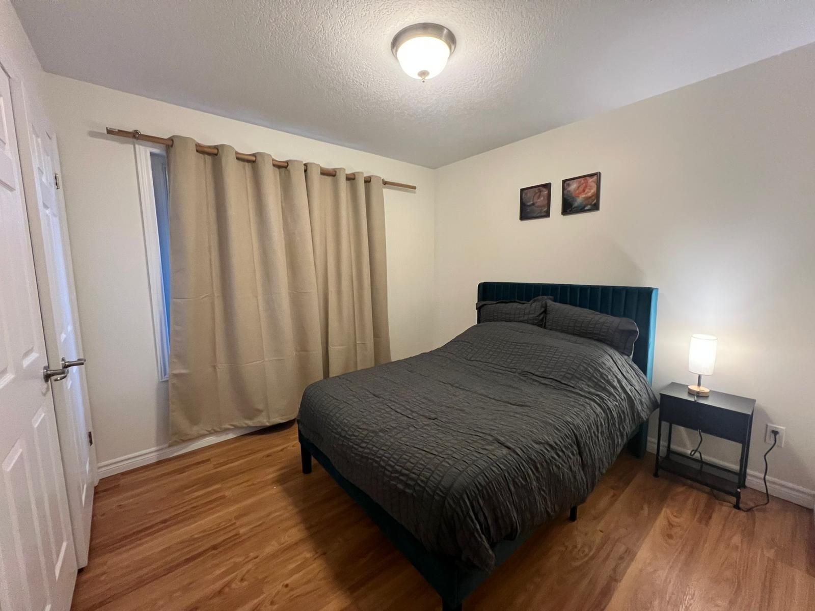 Ein Schlafzimmer mit einem Bett, einem Nachttisch, einer Lampe, einer Deckenleuchte, einer Tür und einem Fenster mit Vorhängen.