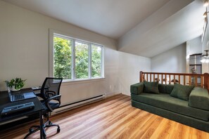 Ein Zimmer mit Holzboden, einem grünen Sofa, einem Schreibtisch mit einem Laptop und einem Fenster mit Blick auf Bäume.