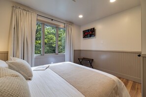 Ein Schlafzimmer mit Bett, einem Fenster mit Vorhängen, einem an der Wand befestigten Fernseher und einem kleinen Tisch.