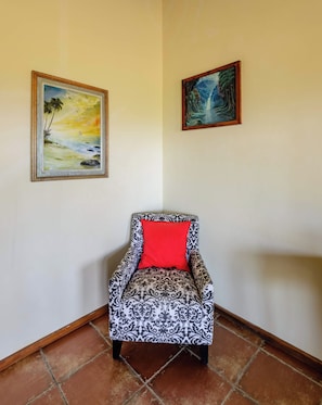 A corner room with a patterned armchair, a red cushion, and two framed paintings on the walls.