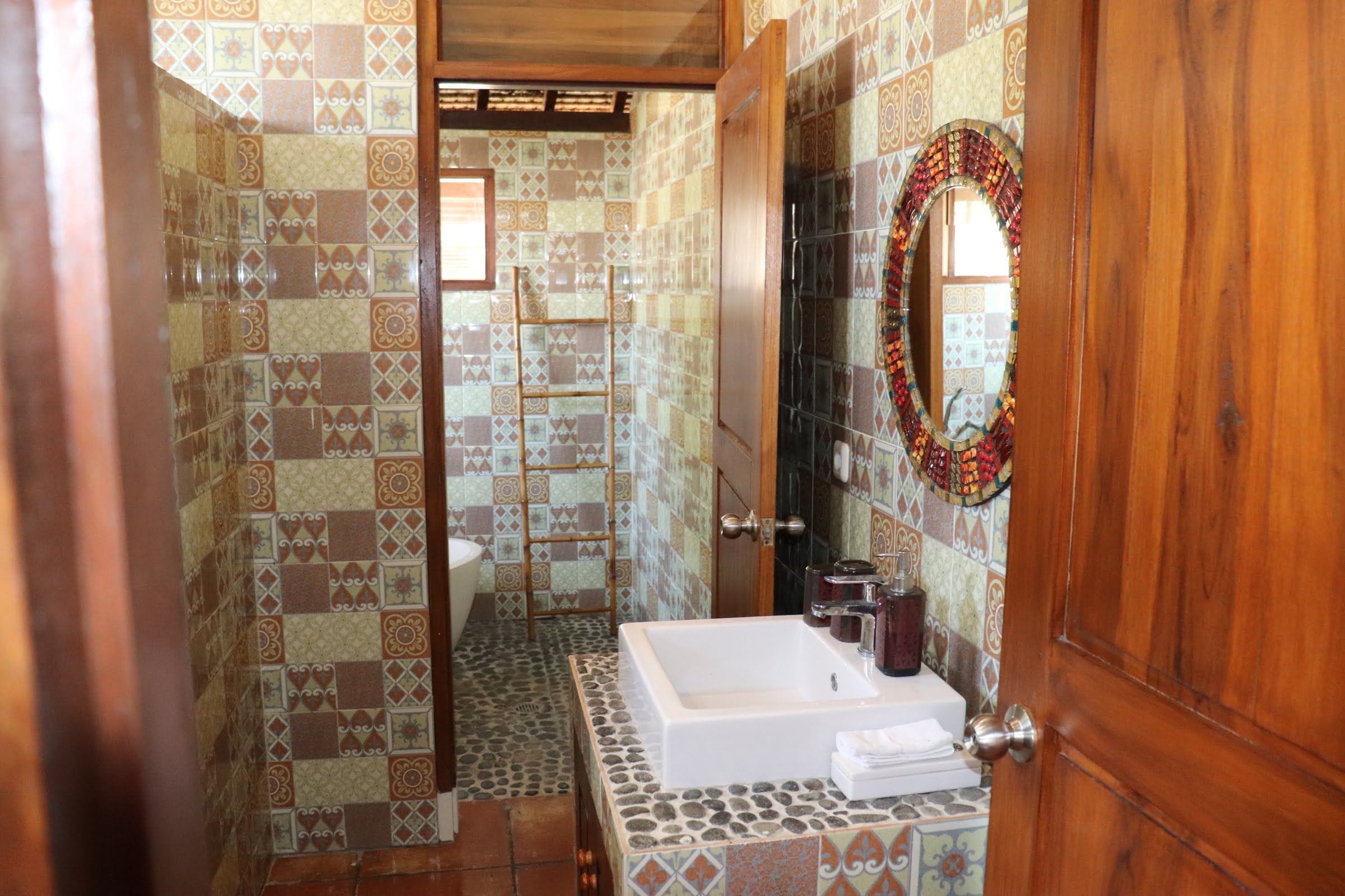 A bathroom with a tiled floor, a white bathtub, a wooden door, and a mirror with a decorative frame.