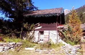 Un edificio rústico de piedra con techo inclinado, rodeado de vegetación natural y un muro de piedra.
