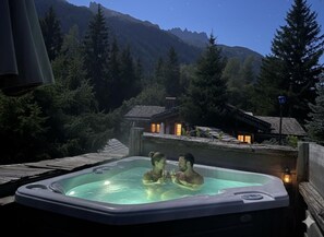 Una vasca idromassaggio con due persone, circondata da un paesaggio montuoso e un pontile in legno.