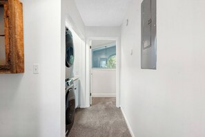 A narrow hallway with a washing machine, a door leading to a room with a window, and a wall-mounted electrical box. - Anastasia Oaks 206B High Tide (St. Augustine)