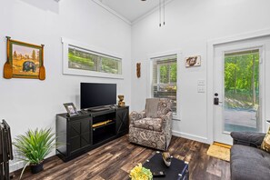 A living room with a TV, a patterned armchair, a sofa, and a large window with blinds. - A Tiny Slice of Paradise (Bryson City)
