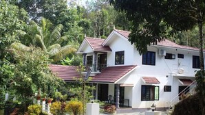 A two-story house with a red-tiled roof, surrounded by lush greenery and a white fence.