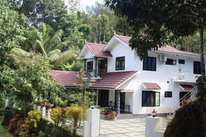 A two-story house with a red-tiled roof, surrounded by lush greenery and a white fence.