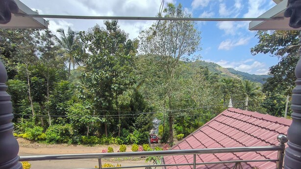 A balcony with a metal railing overlooking a road, surrounded by lush greenery and a red-roofed building.