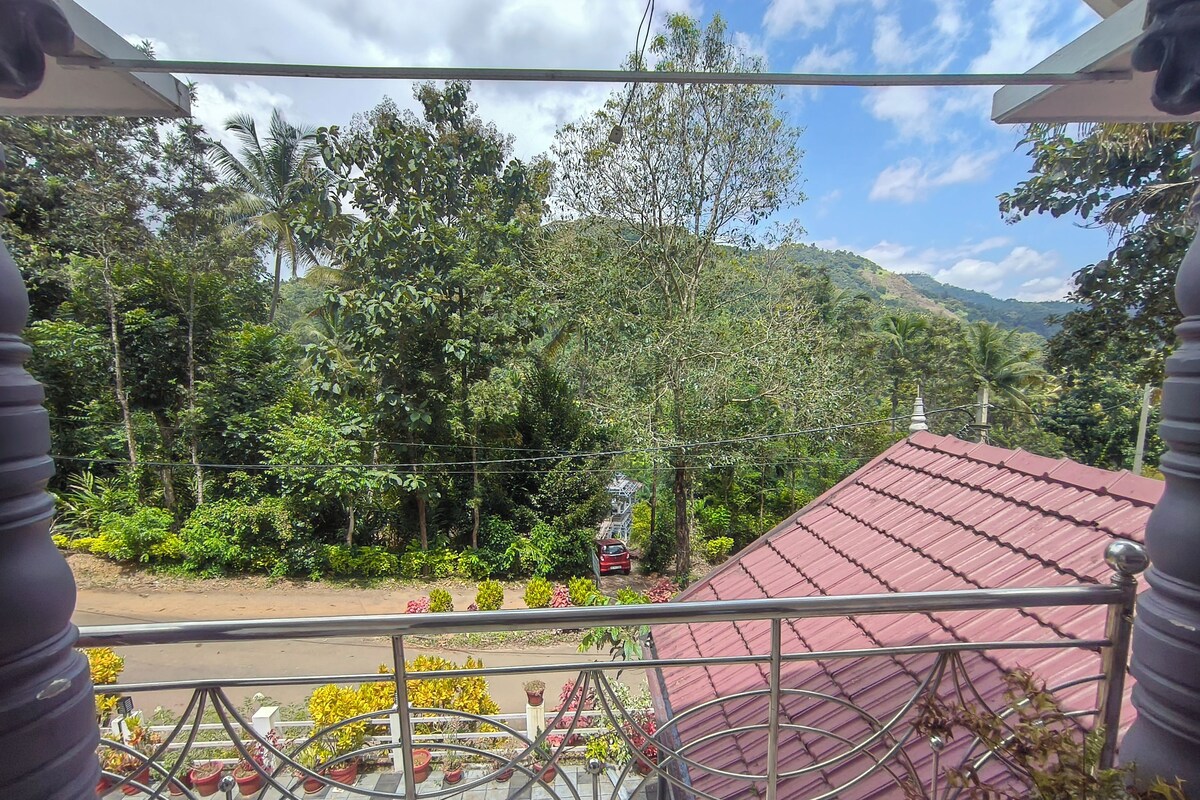 A balcony with a metal railing overlooking a road, surrounded by lush greenery and a red-roofed building.