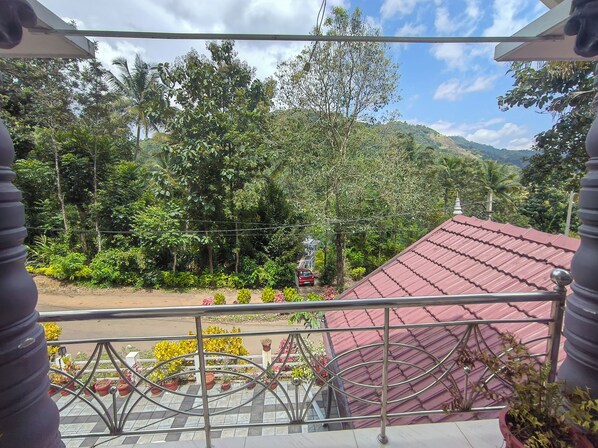 A balcony with a metal railing overlooking a road, surrounded by lush greenery and a red-roofed building.