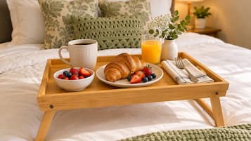 Bandeja de café da manhã com croissants, morangos, amoras, um copo de suco de laranja e uma xícara de café sobre uma cama com travesseiros e uma mesa de cabeceira com abajur.