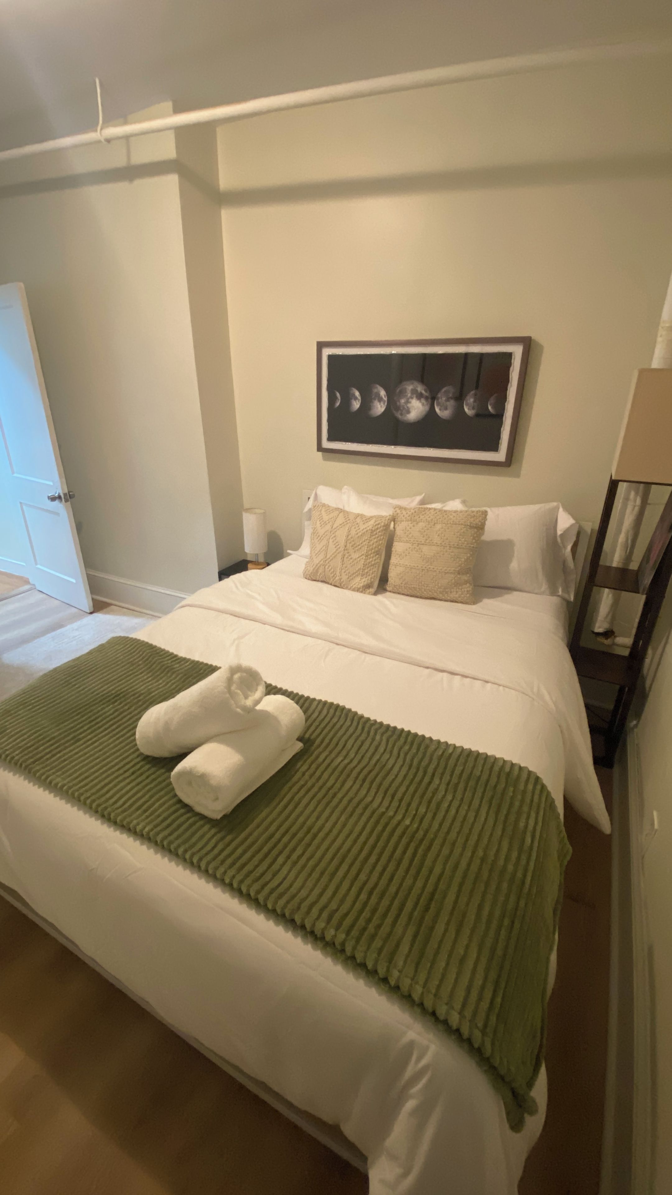 A hotel room with a neatly made bed, a green throw blanket, and a framed picture of the moon phases on the wall.