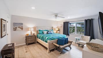 Ein Schlafzimmer mit einem Bett, Nachttischen, einem Sessel, einem kleinen Tisch, einem Deckenventilator und einem Fenster mit Vorhängen.