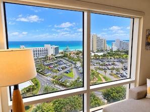 A room with a large window offering a view of a parking lot, a hotel building, and a beach. - Luau 2–6933 | 9th Floor Gulf Views | King + Sleeper Sofa | Tram Included (Miramar Beach)