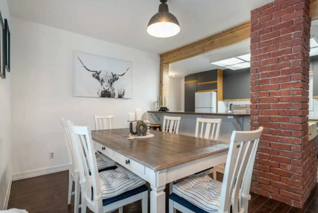 Un coin repas avec une table en bois et des chaises blanches, une cuisine en arrière-plan et une colonne en briques.