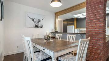Un coin repas avec une table en bois et des chaises blanches, une cuisine en arrière-plan et une colonne en briques.