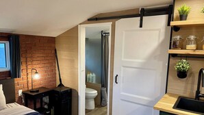 Une chambre à coucher de taille modeste, comprenant un lit, une table de chevet et une lampe. À côté se trouve une salle de bain avec des toilettes et une douche. On aperçoit également un coin cuisine avec un évier et des étagères.