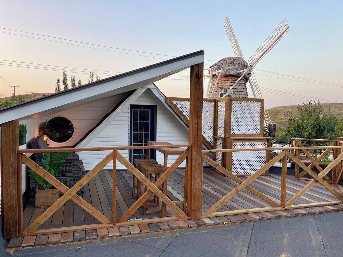 Une terrasse en bois avec un moulin à vent en arrière-plan, une petite maison avec un toit en pente et une clôture en bois.