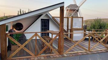 Une terrasse en bois avec un moulin à vent en arrière-plan, une petite maison avec un toit en pente et une clôture en bois.