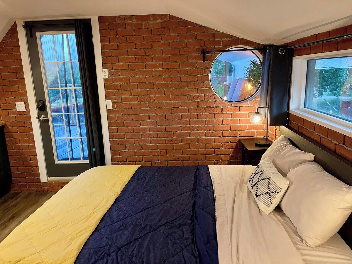 A bedroom with a brick wall, a bed with a blue and white duvet, a round mirror, and a window with a view.